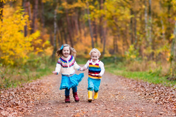 Zwei Kinder in farbenfrohen Kleidern laufen Hand in Hand einen mit Blättern bedeckten Weg in einem lebhaften Herbstwald entlang.